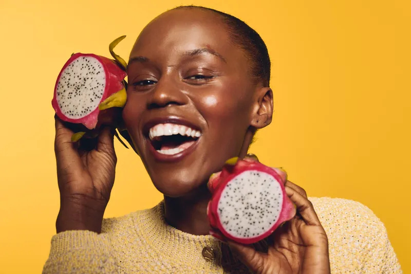Smiling African woman with dark skin holding two halves of dragon fruit near her face on yellow background. Concept of healthy lifestyle, tropical fruit, happiness, and natural beauty.