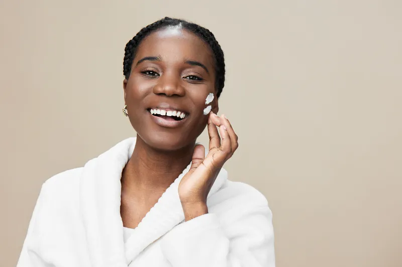 Smiling woman applying skincare cream on her face in a cozy white robe, highlighting healthy skincare and natural beauty against a warm, neutral background