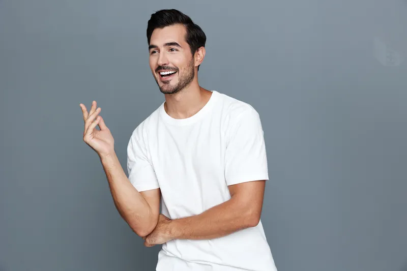 Full body smiling happy cheerful young man wearing a white t-shirt poses confidently against a solid gray background, expressing joy and warmth in a bright studio setting.