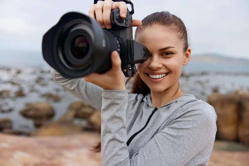 Young woman smiling while holding a camera on the beach, showcasing passion for photography and outdoor adventures in a casual gray outfit