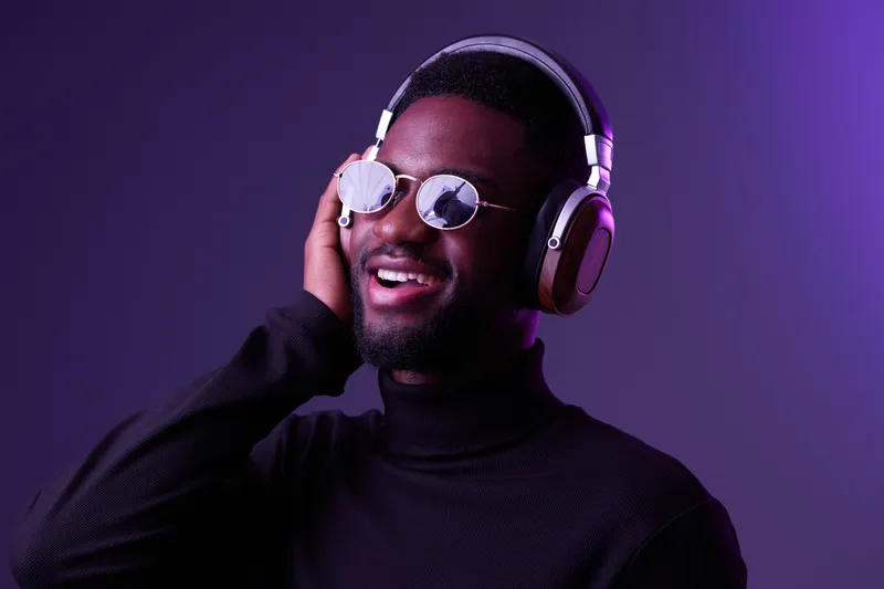 Happy young Black man enjoying music with headphones, wearing stylish sunglasses against a vibrant purple background, ideal for music and lifestyle themes