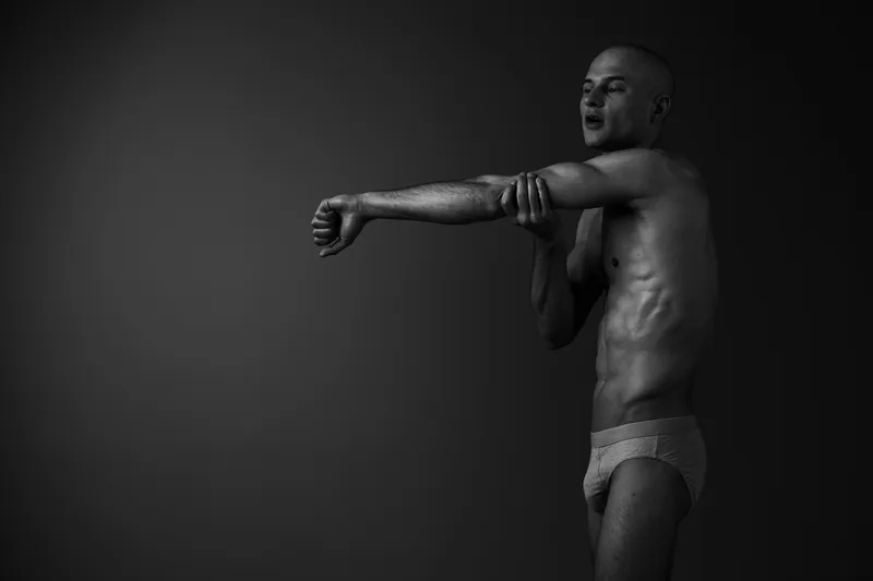Black and white image of a man stretching his arms, showcasing a fit physique and healthy posture in a minimalist setting. A focus on body movement and form.