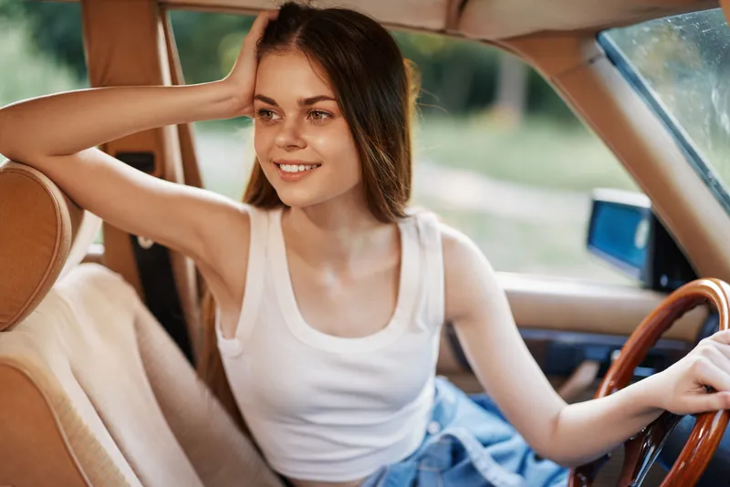 Young woman smiling in a vintage car, wearing a casual white tank top and denim shorts, surrounded by a natural green background, conveying a carefree summer vibe.