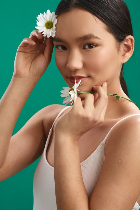 asian woman with a flower tucked behind her ear, holding a stem near her lips, posed against a vibrant green backdrop to convey fresh beauty and serene elegance.
