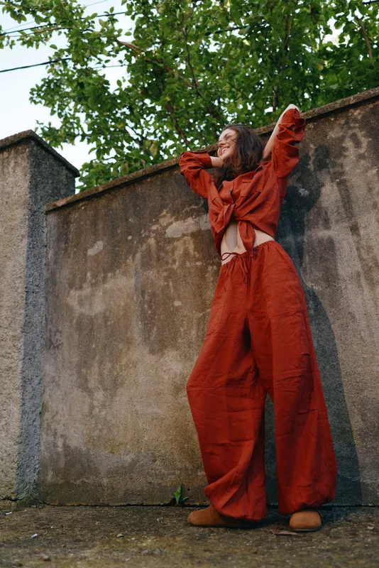 Young woman in an orange wideleg jumpsuit stretches against a weathered wall outdoors, striking a bold pose with a confident expression and a modern urban vibe