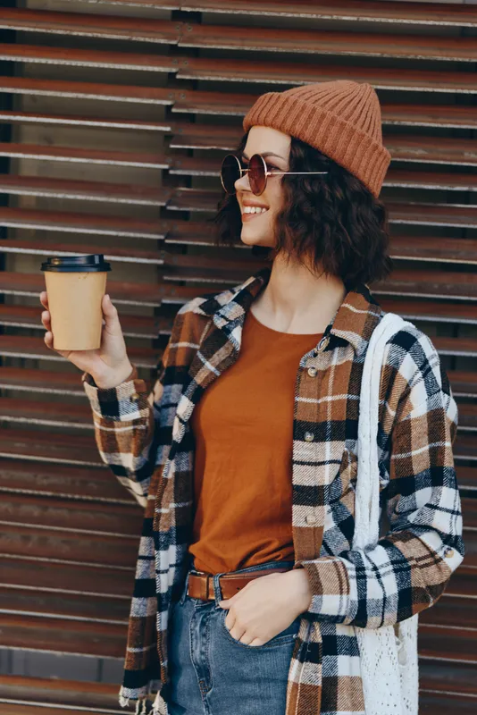 Woman holding coffee and smile in beanie and sunglasses, plaid shirt and tote bag in urban setting. Candid lifestyle image with authenticity, golden hour glow and mindful living vibe.