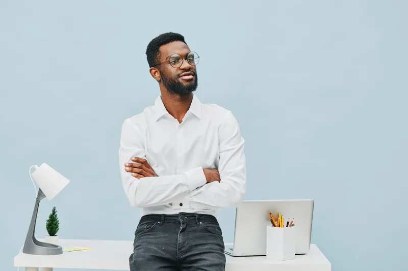 Professional man with glasses wearing white shirt standing near workspace with laptop and stationery isolated on light blue background. People business workplace concept