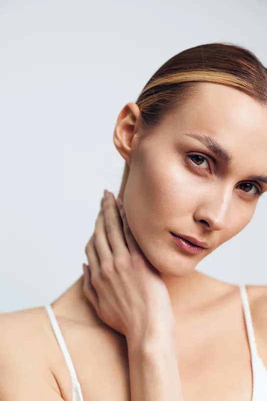 Close-up portrait of a young woman with clear skin touching her neck, looking at the camera, minimal makeup, soft lighting, neutral background, concept of skincare and beauty.