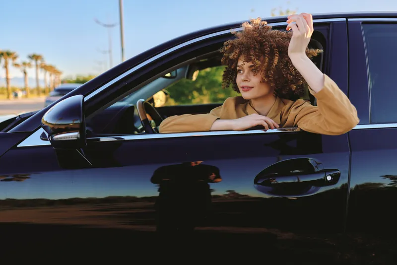 Young woman with curly brown hair leans out of a sleek black car window, smiling softly as a warm breeze enhances the sunny day and creates a relaxed candid moment for a lifestyle shoot