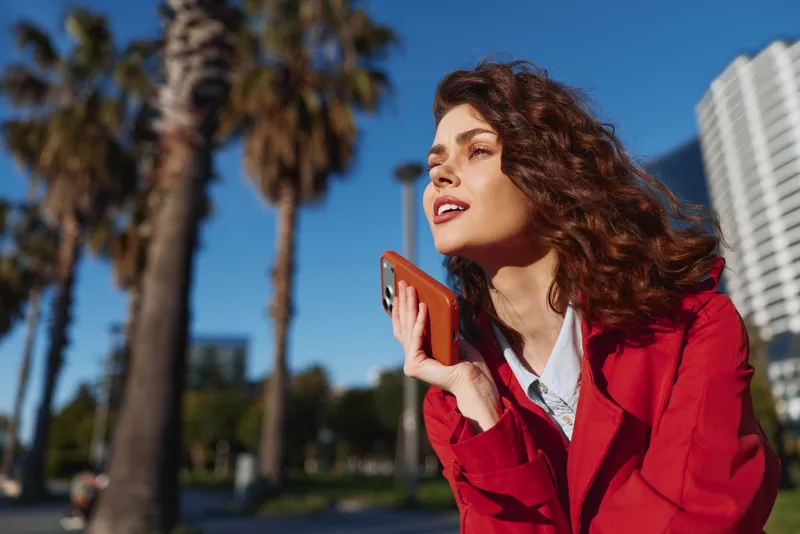A young woman with curly hair wearing a red coat holds a smartphone close to her face outdoors with palm trees and buildings in the background on a sunny day.