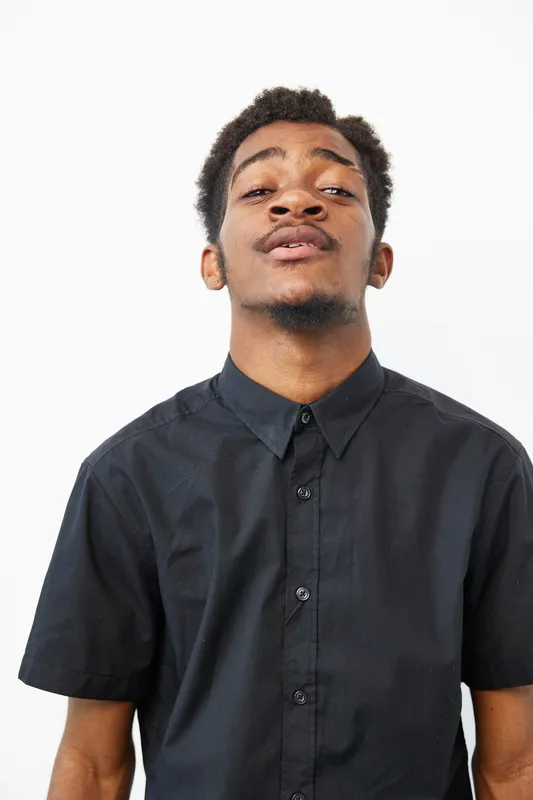 A confident black man in a black shirt exudes charisma and poise, showcasing his unique style. This vibrant portrait captures individuality and strength, reflecting urban fashion.