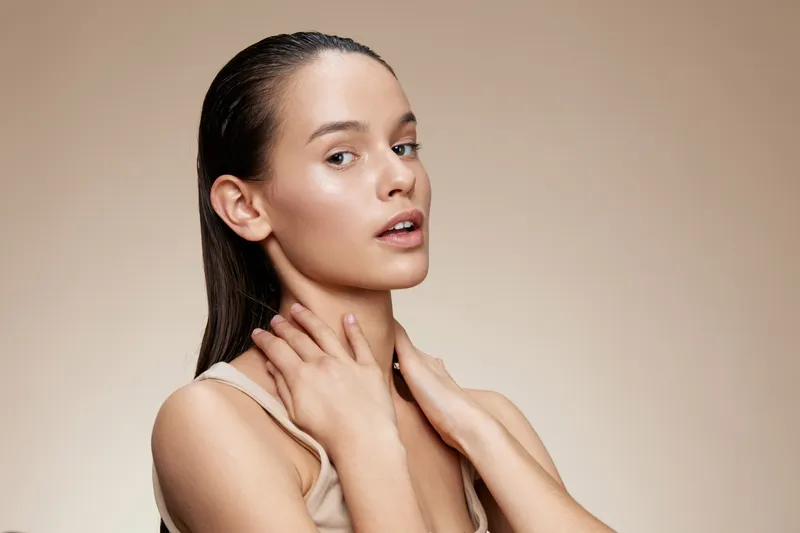 A serene young woman with long hair in a neutral toned outfit gently touching her neck, showcasing natural beauty against a soft beige background, emphasizing confidence and elegance