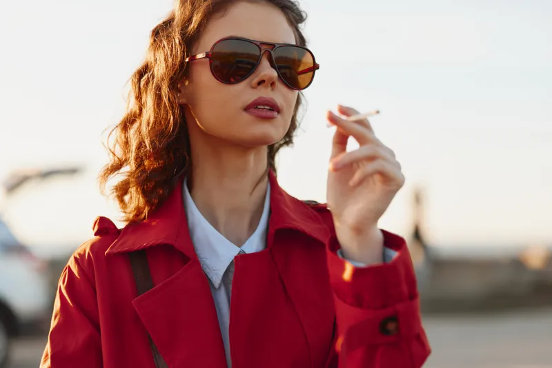 Young woman in a red coat and sunglasses smokes outdoors, sunlight glow, confident pose in an urban setting.