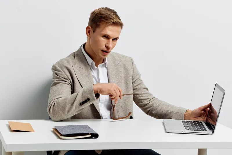 Professional young man in smart casual attire using laptop and holding eyeglasses while working at a desk isolated on plain white background. Office, business, startup, technology, communication conce