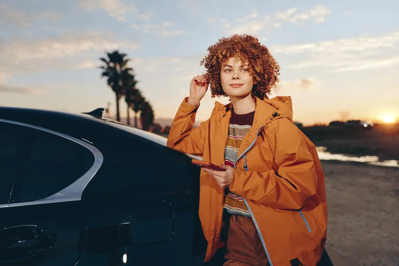 Woman lifestyle car sunset jacket outdoors travel road curly hair, a confident woman in a brown coat stands beside a parked car, touching her hair while preparing to leave at sunset on open road
