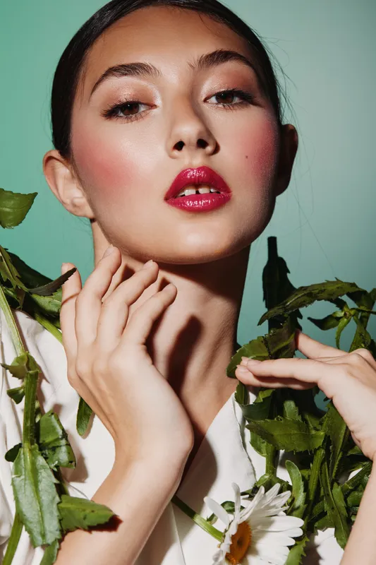 Portrait of a young woman with glossy red lips and flushed cheeks posing with greenery against a soft green background. Creative beauty and fashion concept emphasizing bold makeup.