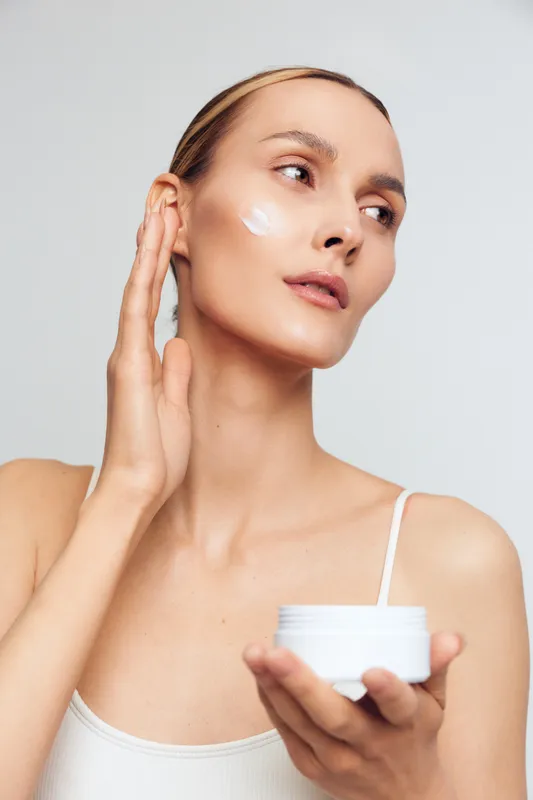 Smiling woman applying face cream on cheek, holding a white jar. Close up on light gray background, soft studio lighting. Skincare self care wellness concept, daily moisturizer