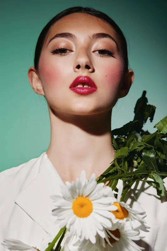 Fashion model with glossy red lips and strong blush poses with white daisies on green background. Creative beauty portrait showcases fresh floral style concept.