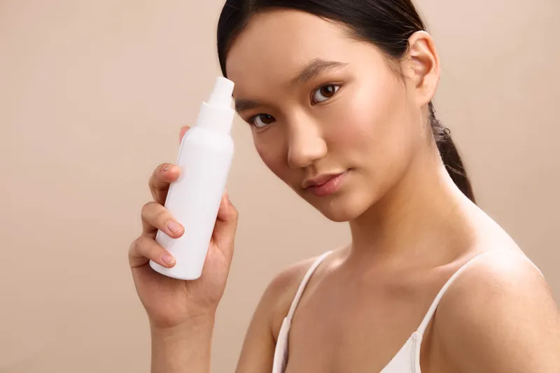 asian woman applies skincare product softly on cheek in a clean studio, exuding calm confidence and radiant skin, creating an inviting beauty moment focused on care and balance.
