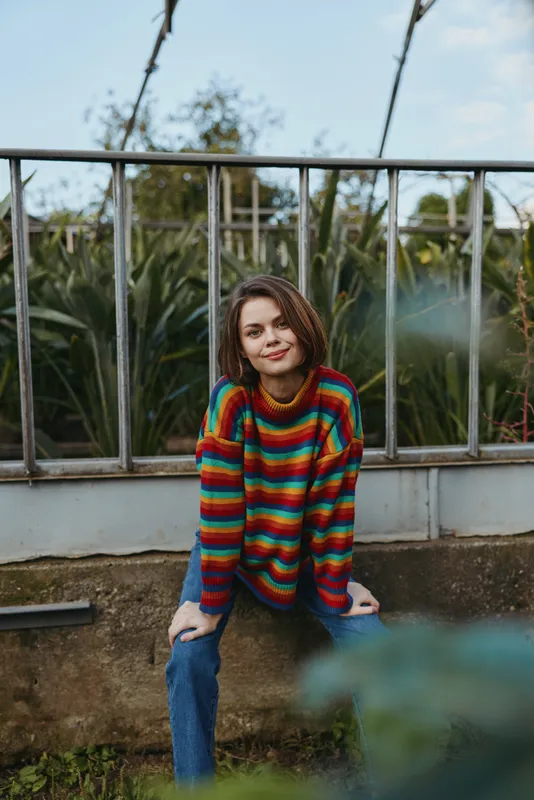 Woman portrait in colorful rainbow sweater, casual outdoor scene with braids and relaxed smile seated by railing in garden with natural light and cozy style