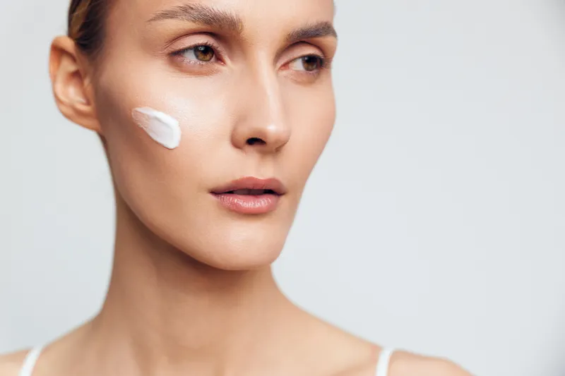 Young woman with clear skin applies white cream on cheek looking away thoughtful isolated on white background closeup beauty skincare concept.