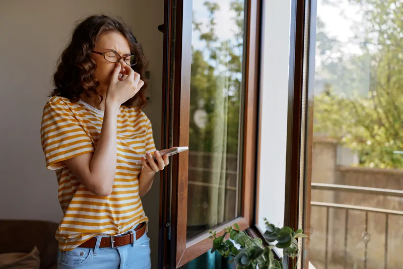 Smiling woman with striped shirt standing by a sunny window, checking her phone in a cozy home setting. Casual lifestyle scene focused on daily routine, comfort, and selfcare. Warm natural light