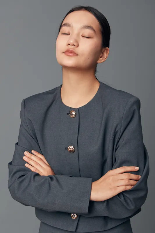 Woman with crossed arms wearing gray jacket with closed eyes isolated on gray background showing confidence and calmness with relaxed posture and neat hairstyle.