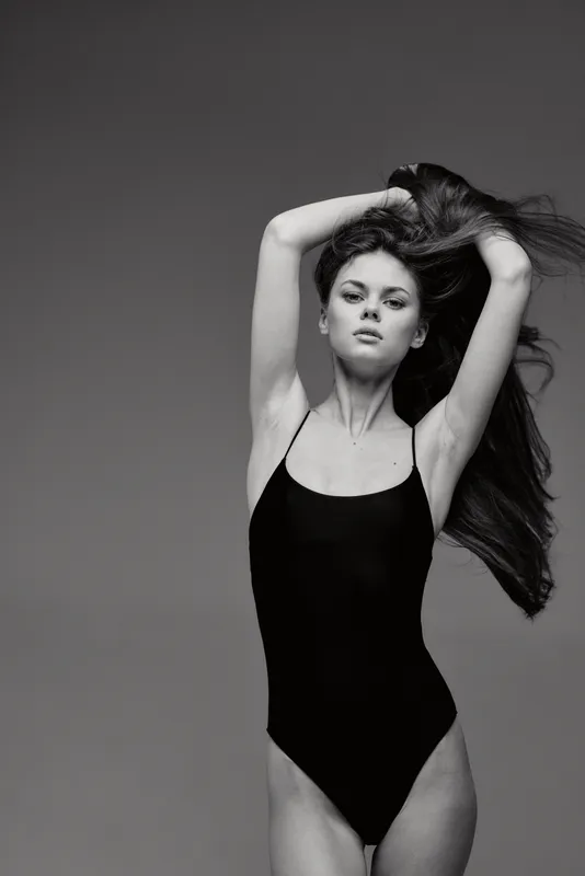 Black and white portrait of a confident woman posing in a black bodysuit with flowing hair and lifted arms