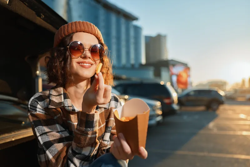 woman eating fries by car in parking lot at golden hour, smiling in sunglasses and beanie; candid lifestyle authenticity, golden hour glow and mindful living in a genuine urban moment.