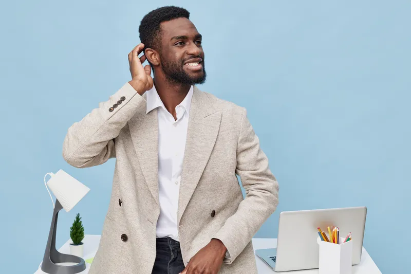 Confident smiling man with dark skin and curly hair wearing beige blazer and white shirt, standing at desk with modern laptop, desk lamp, and stationery on blue background. Business and technology con