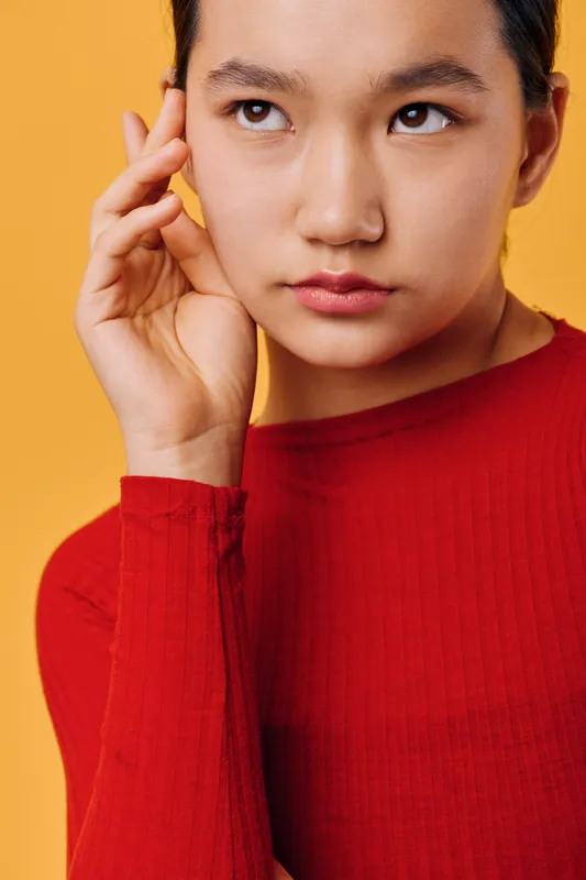 Young woman with clear skin and dark hair wears red sweater touching her face on warm orange background. Beauty portrait with serious expression and thoughtful look, closeup in studio.
