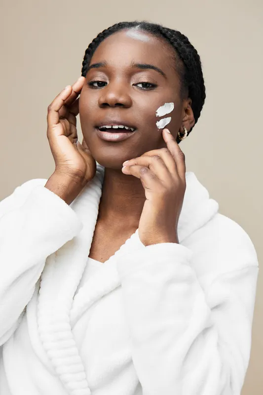 Young African woman applying facial cream with a joyful expression in a light colored background, emphasizing skin care and beauty routines