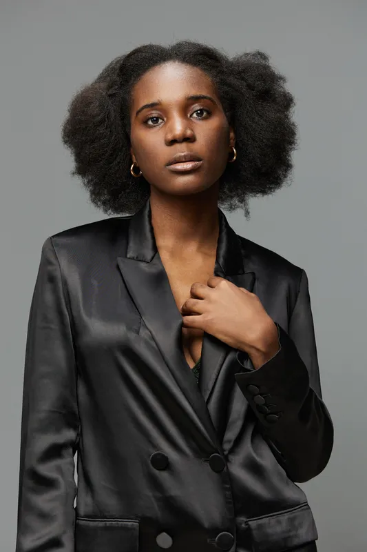 Elegant portrait of a woman with natural hair wearing a black satin blazer, expressing confidence against a neutral background