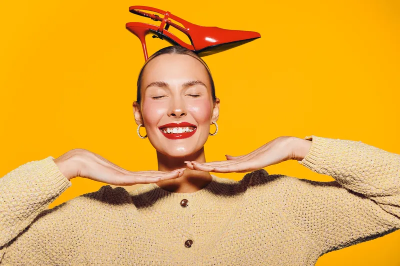 Smiling woman with closed eyes balances red high heel shoes on her head against bright yellow background. Concept of fun, fashion, joy, and playful mood in studio portrait.
