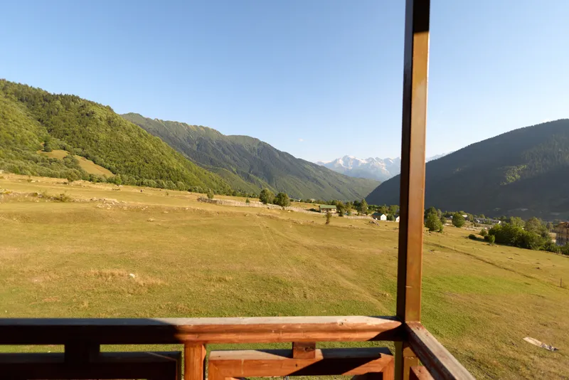Balcony wooden railing overlooking meadow and mountains in a wide valley landscape. Sunny sky, green hills and grass fields with distant peaks and peaceful rural view.