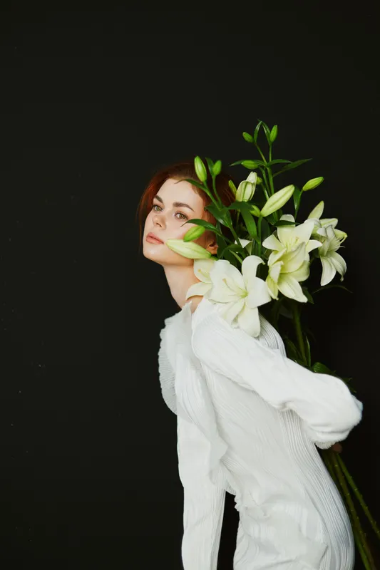Young woman in a white dress holding white lilies against a dark background, conveying elegance and serenity in a spring floral concept