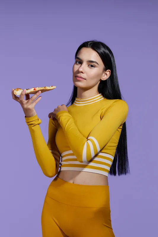Woman sport fitness dressed in yellow activewear holds a slice of pizza against a purple background, showcasing a blend of health and casual indulgence with a confident expression.