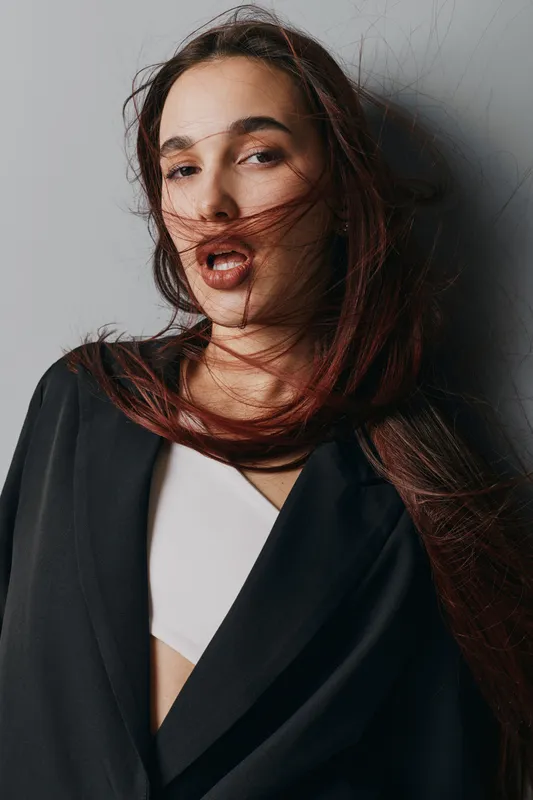 Woman with long hair blowing in wind wearing black blazer and white top standing against gray background with confident expression and natural makeup in studio portrait.