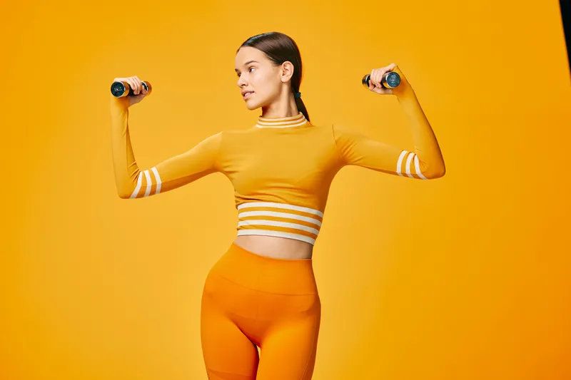 Fitness themed portrait of a woman in orange activewear lifting dumbbells with a bold orange background, showcasing strength, balance and athletic determination in a dynamic studio.