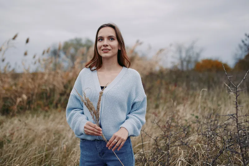 Young woman standing in a windswept field holding dried grasses, a natural candid portrait conveying authenticity and grounded presence, gentle expression and realistic lifestyle details.
