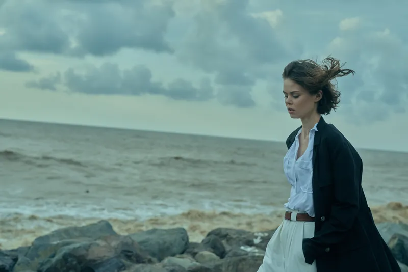 Woman on a rocky shoreline, wind in her hair, wearing a white shirt and dark jacket, gazing toward the sea beneath moody clouds, a fashion mood by the coast