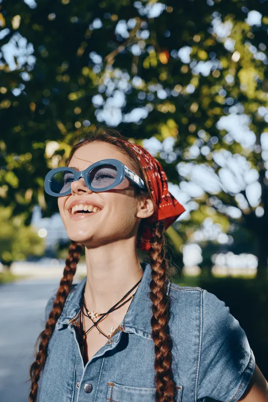 smiling woman with braided hair wearing blue sunglasses and denim jacket outdoors under bright sunlight in summer day