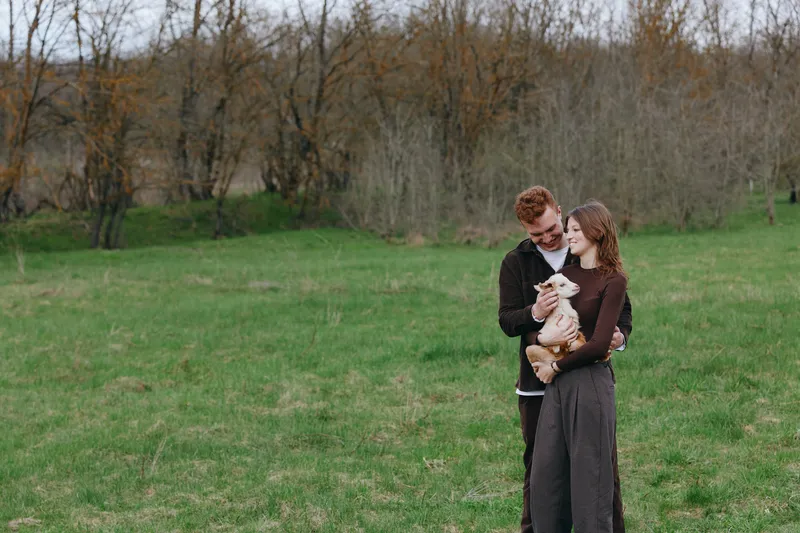 A couple outdoors in a field share a tender moment with a goat kid, smiling as they connect with their animal. A lifestyle portrait of affection, rural living, and simple bonding in daylight.