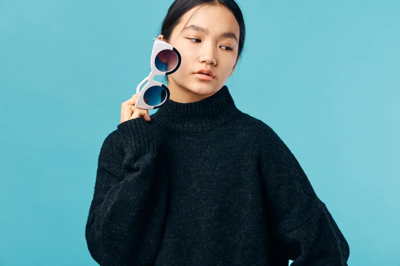 Young woman in dark sweater holding sunglasses near face with neutral expression on blue background. Casual style with minimal makeup and contemplative mood for fashion or lifestyle concept.