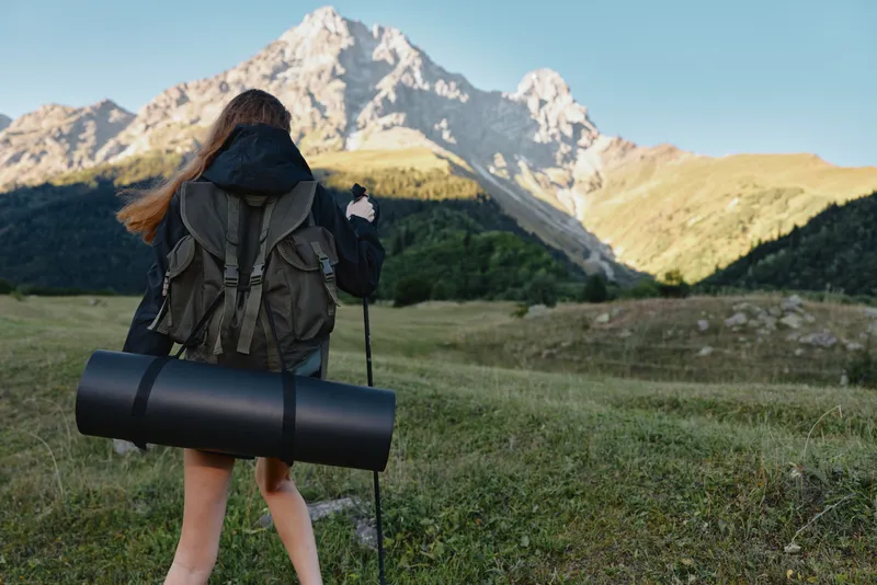 A female hiker walks through a grassy meadow toward rugged mountains, carrying a rolled sleeping mat and a backpack, bathed in clear outdoor light.