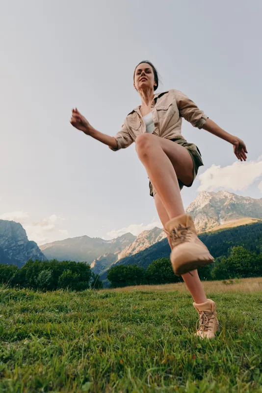 Active outdoor woman jumps across a grassy field with mountains behind, capturing energetic lifestyle moments, sunlit scene, vibrant mood, and a sense of freedom today