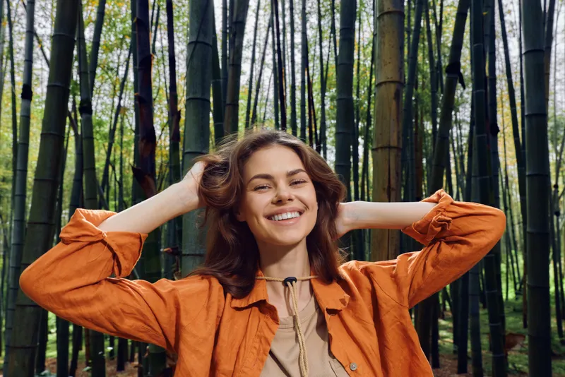 Woman smile bamboo forest portrait outdoors nature happy in orange jacket, young female relaxed and smiling among tall bamboo stalks, serene lifestyle and natural beauty expression.