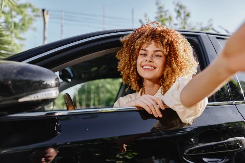 smiling woman with curly hair sitting in black car, offering hand outside window, casual clothing and natural light in outdoor setting, happy expression