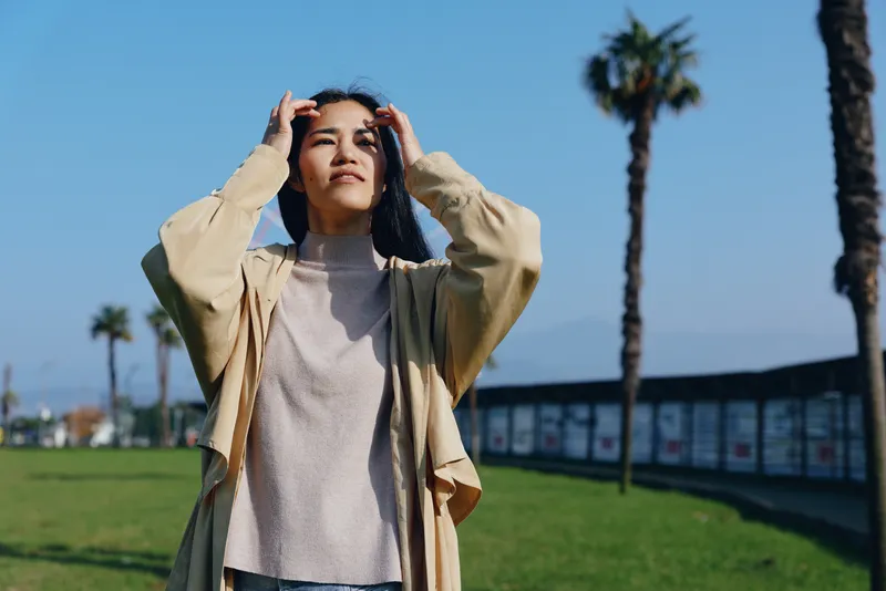 Outdoor woman casual summer park sunshine palm vibe with relaxed pose as she raises hands to adjust hair blue sky green grass background light cardigan and sweater candid lifestyle scene in a sunny