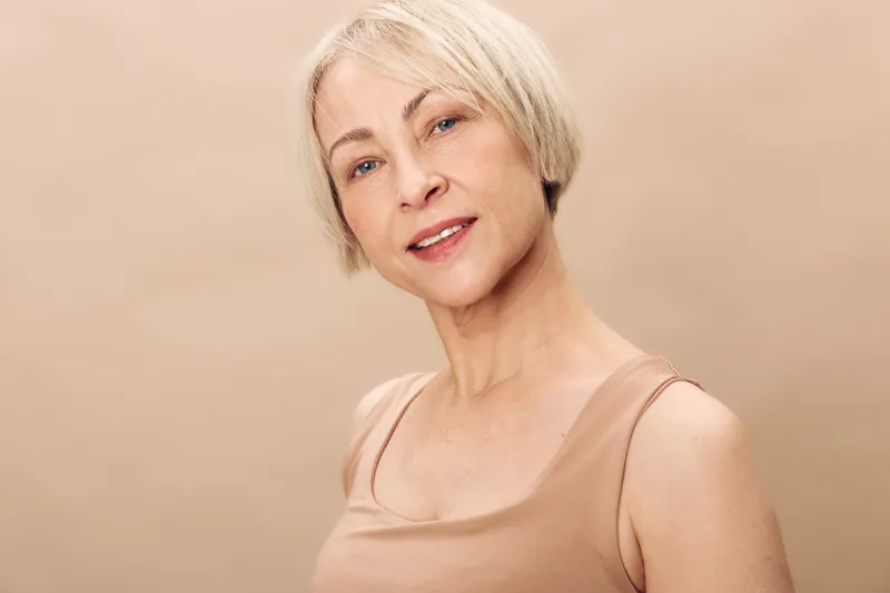 Portrait of a mature woman with short blonde hair smiling gently, wearing a beige tank top against a neutral background. Concept for aging beauty, confidence, and natural skin care.